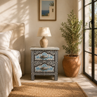 Lifestyle scene with a hand-painted nightstand made from reclaimed wood, featuring floral motifs in soft turquoise and black with intricate carved detailing and two drawers for storage.

