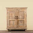 A rustic carved wooden cabinet with two doors, featuring intricate floral and oval patterns on the doors, black metal handles, and turned wooden feet, set against a plain beige wall.