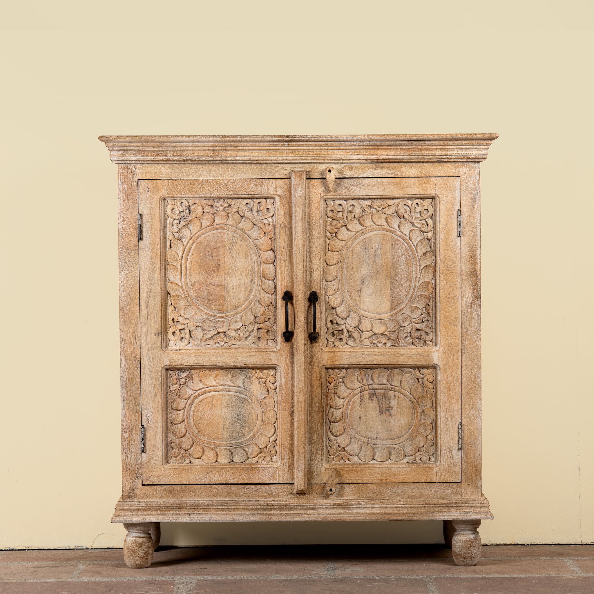 A rustic carved wooden cabinet with two doors, featuring intricate floral and oval patterns on the doors, black metal handles, and turned wooden feet, set against a plain beige wall.