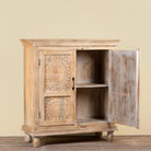 A small carved wooden cabinet with one door open, showing an interior shelf. The cabinet has ornate floral and circular carvings on the left door and a distressed, weathered finish.