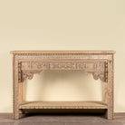 An ornate carved wooden console table with a natural light wood finish, featuring intricate floral carvings on the apron, legs, and lower shelf, set against a plain beige wall.