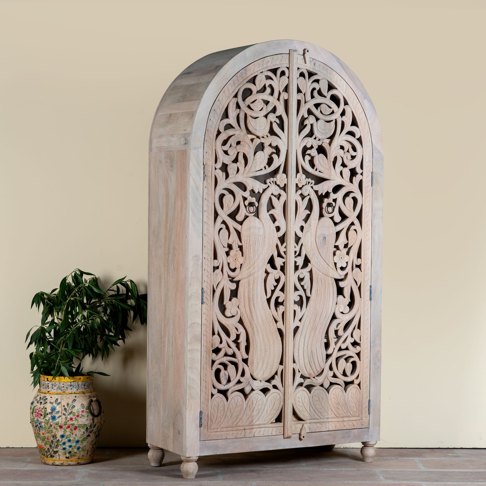 Side angle image of a hand-carved mango wood wardrobe with arched top and intricate peacock floral design – artisanal Indian bedroom furniture.