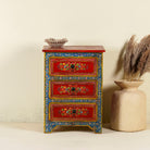 A hand-painted reclaimed wood nightstand with vibrant floral motifs, featuring three drawers and a recycled wood top, placed next to a vase with dried plants.