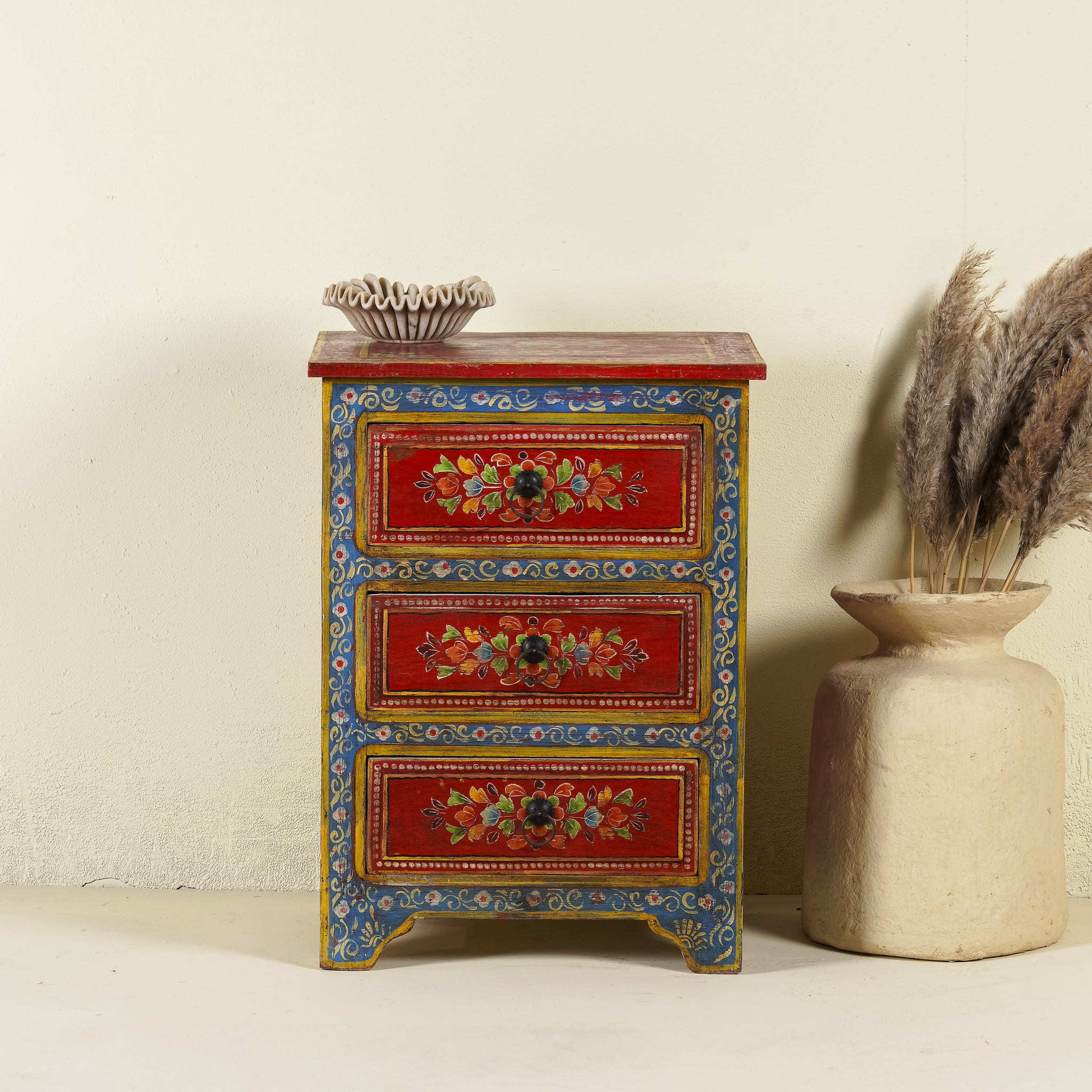 A hand-painted reclaimed wood nightstand with vibrant floral motifs, featuring three drawers and a recycled wood top, placed next to a vase with dried plants.