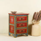 Side angle image of a hand-painted reclaimed wood nightstand with vibrant floral motifs, featuring three drawers and a recycled wood top, placed next to a vase with dried plants.