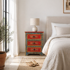 Lifestyle scene with a hand-painted reclaimed wood nightstand with vibrant floral motifs, featuring three drawers and a recycled wood top, placed next to a vase with dried plants.