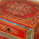 Close up image of a hand-painted reclaimed wood nightstand with vibrant floral motifs, featuring three drawers and a recycled wood top, placed next to a vase with dried plants.