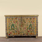 An antique wooden chest or sideboard with intricate floral tapestry panels, featuring a beige background with colorful embroidered flowers, leaves, and birds, set against a plain beige wall and rustic wooden floor.