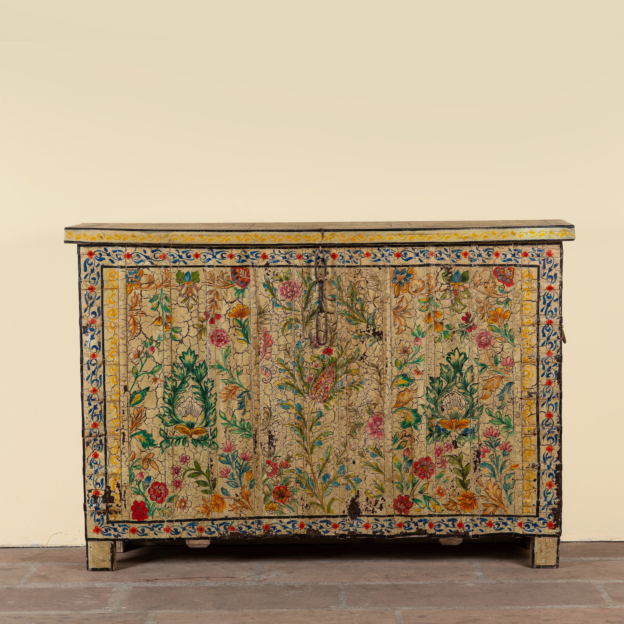 An antique wooden chest or sideboard with intricate floral tapestry panels, featuring a beige background with colorful embroidered flowers, leaves, and birds, set against a plain beige wall and rustic wooden floor.