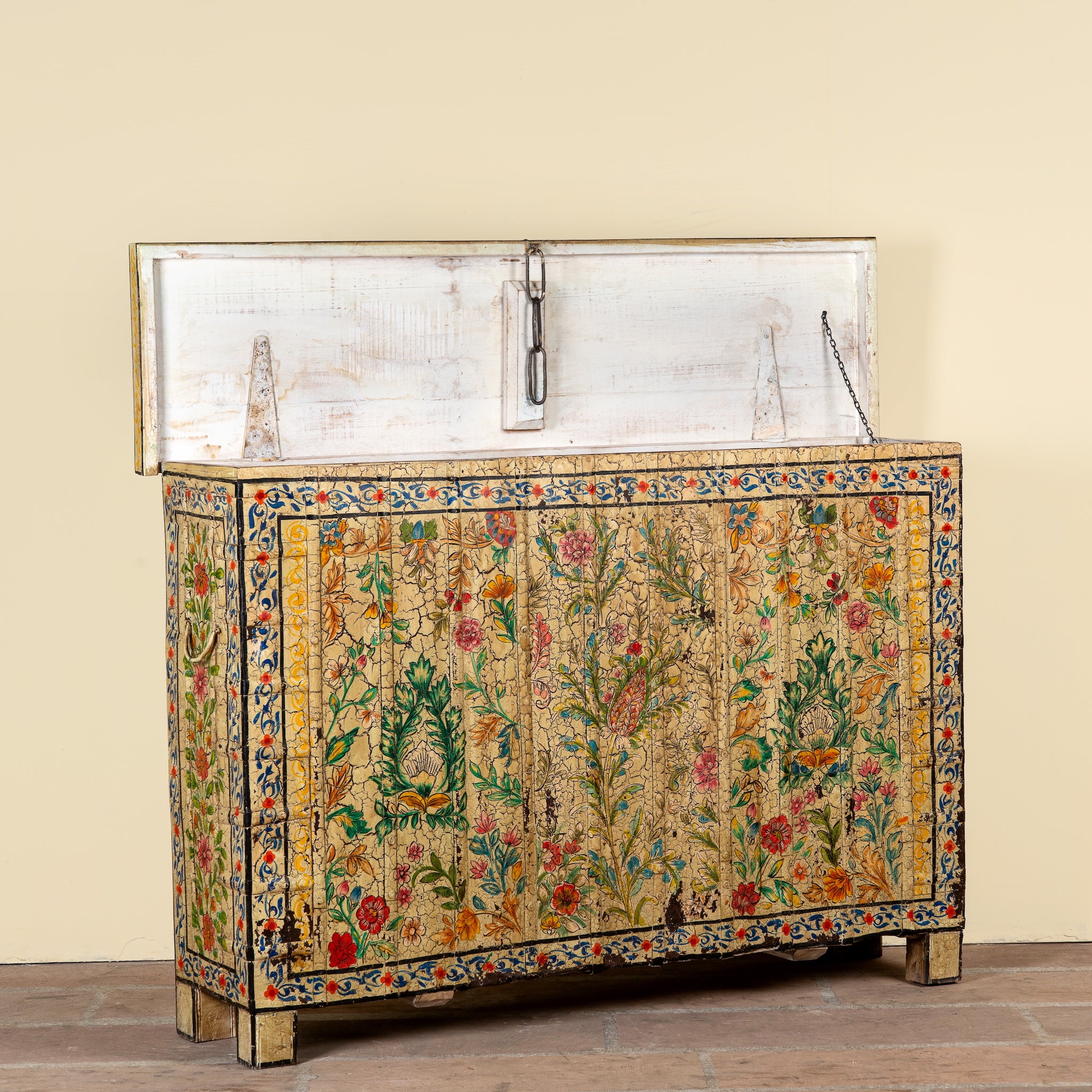 An ornate antique-style wooden chest or cabinet with a distressed white top panel and a heavily decorated front featuring intricate floral and botanical motifs in green, red, orange, and blue on a gold-toned background. The cabinet stands on a wooden floor against a plain beige wall.
