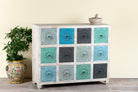 Side angle image of a carved mango wood chest of drawers with twelve multicolour floral drawer fronts, 124 × 40 × 102 cm.