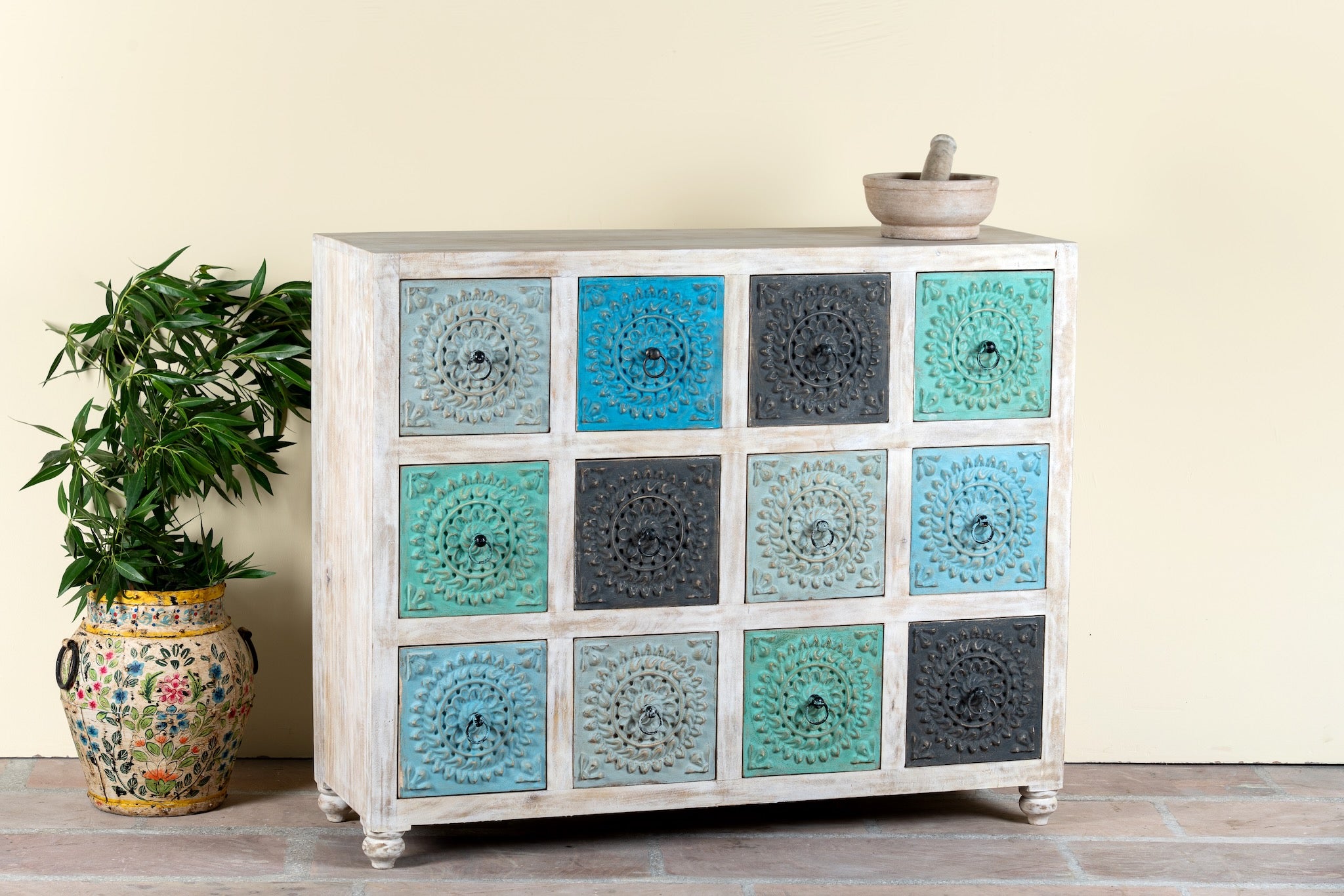 Side angle image of a carved mango wood chest of drawers with twelve multicolour floral drawer fronts, 124 × 40 × 102 cm.