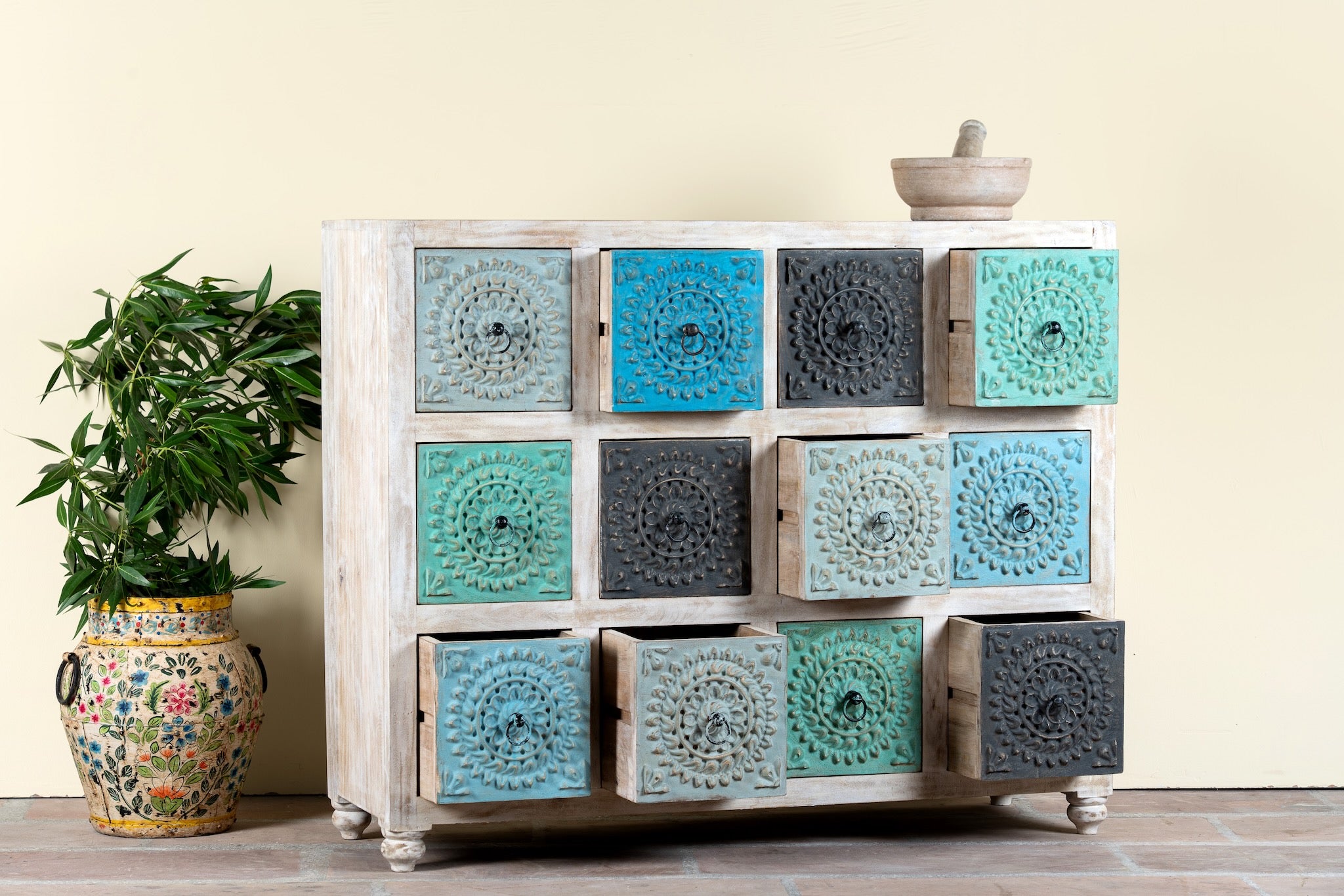 Side angle image with open drawers of a carved mango wood chest of drawers with twelve multicolour floral drawer fronts, 124 × 40 × 102 cm.
