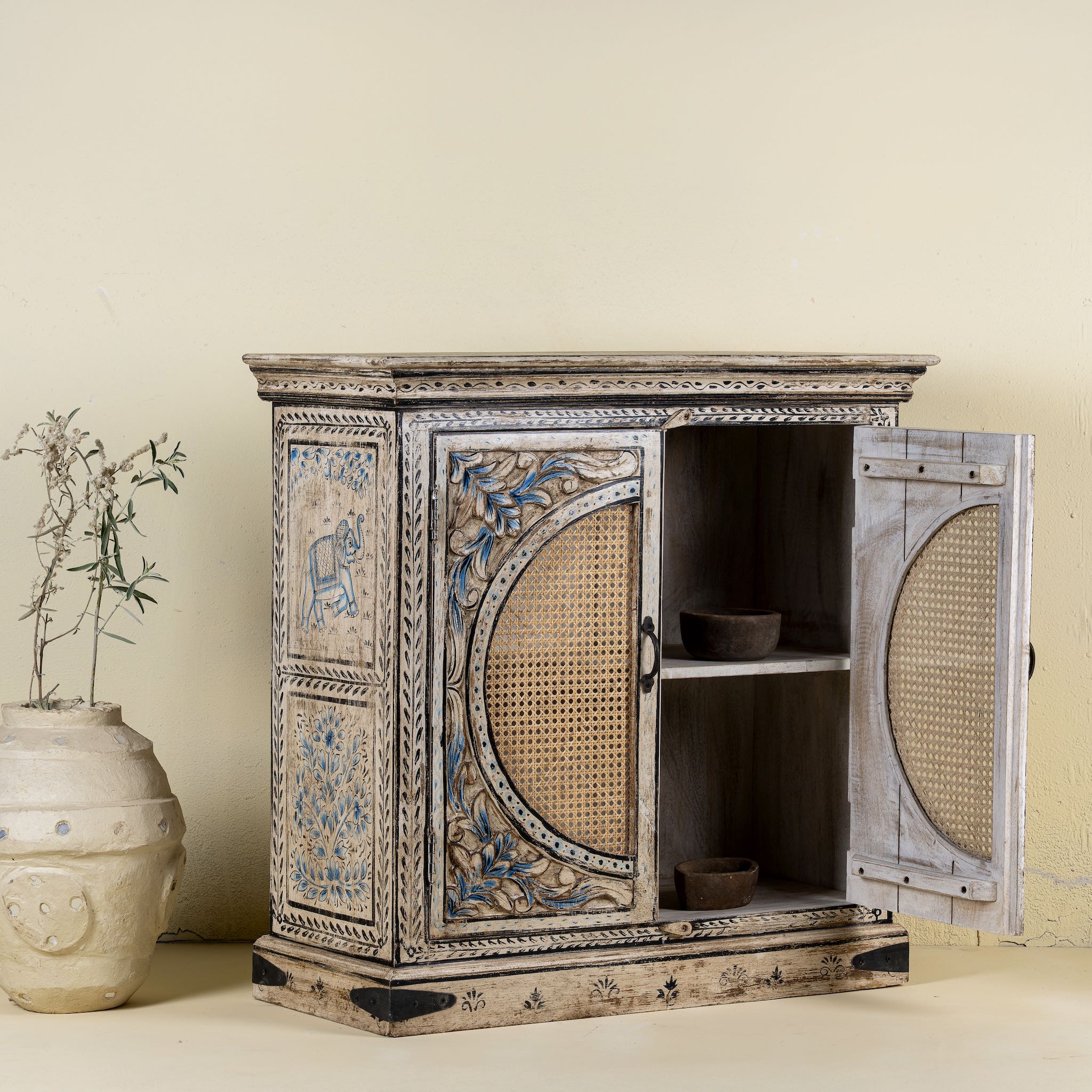 Open door view of a hand-made, hand-carved cupboard made of mango wood with blue floral designs, elephant motifs and rattan inserts.