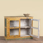 A small hand-painted wooden cabinet with two open doors, showcasing a distressed white wood interior and colorful green, yellow, and red floral borders. A small woven basket sits on top of the cabinet against a solid beige wall.