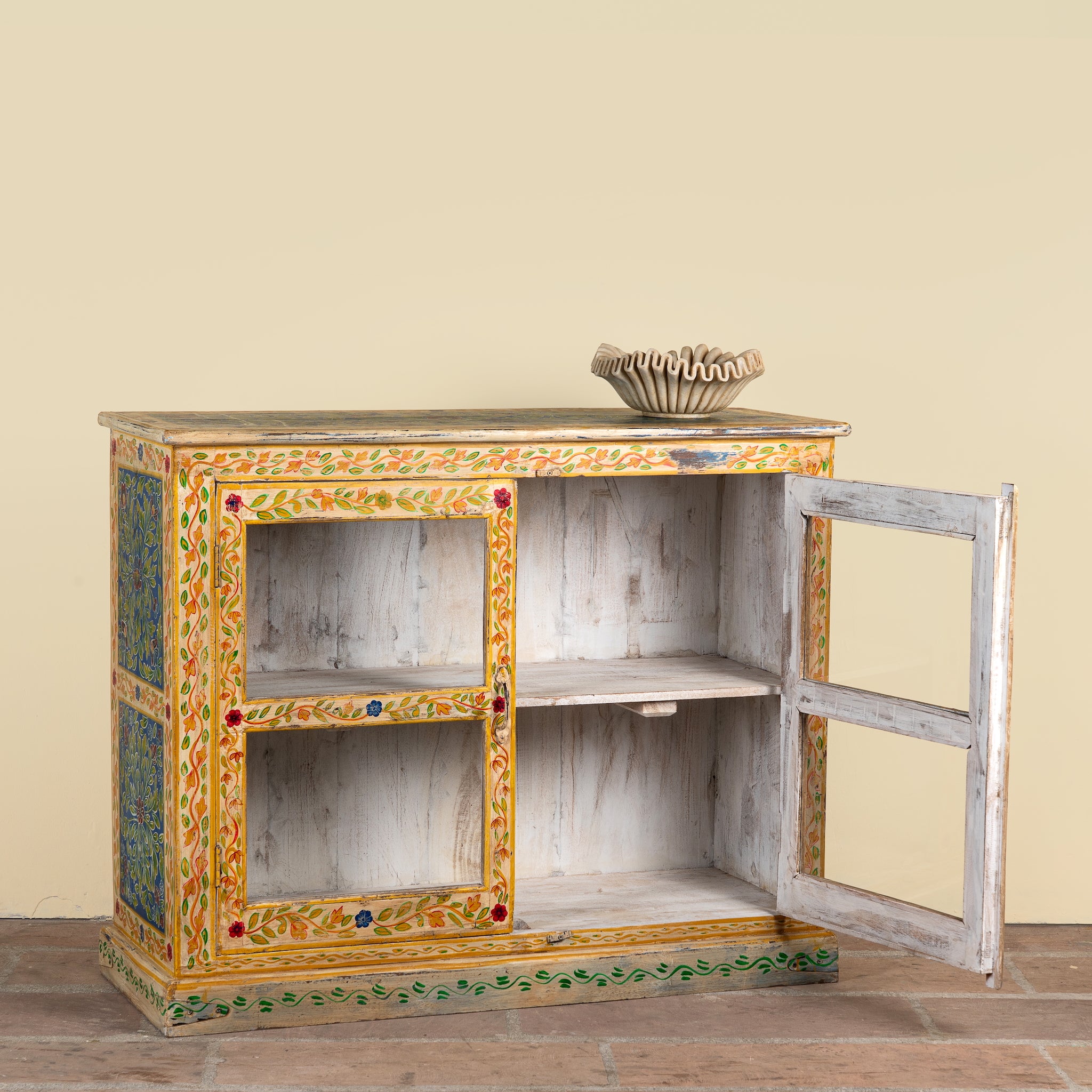 A small hand-painted wooden cabinet with two open doors, showcasing a distressed white wood interior and colorful green, yellow, and red floral borders. A small woven basket sits on top of the cabinet against a solid beige wall.