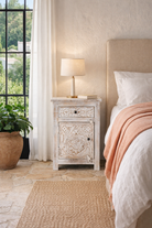 Lifestyle imagination of a pair of mango wood nightstands Uday with whitewashed vintage finish, featuring hand-carved geometric and floral designs. Each bedside table includes a drawer and cabinet for elegant and practical storage.