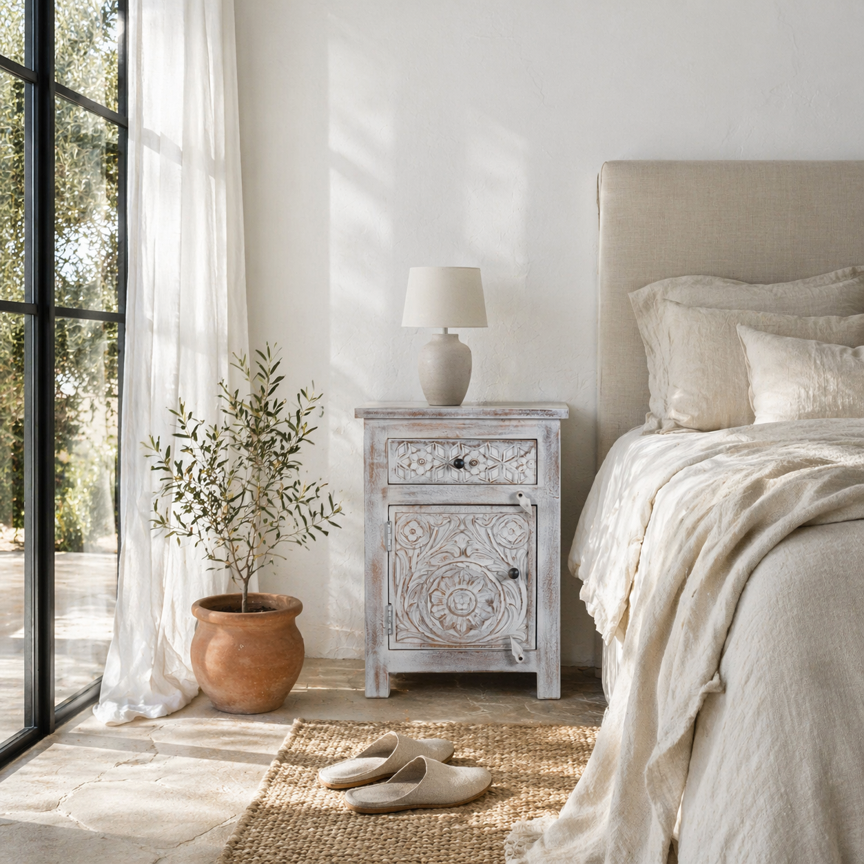 Lifestyle image of a mango wood nightstand Uday with whitewashed vintage finish, featuring hand-carved geometric and floral designs. Each bedside table includes a drawer and cabinet for elegant and practical storage.