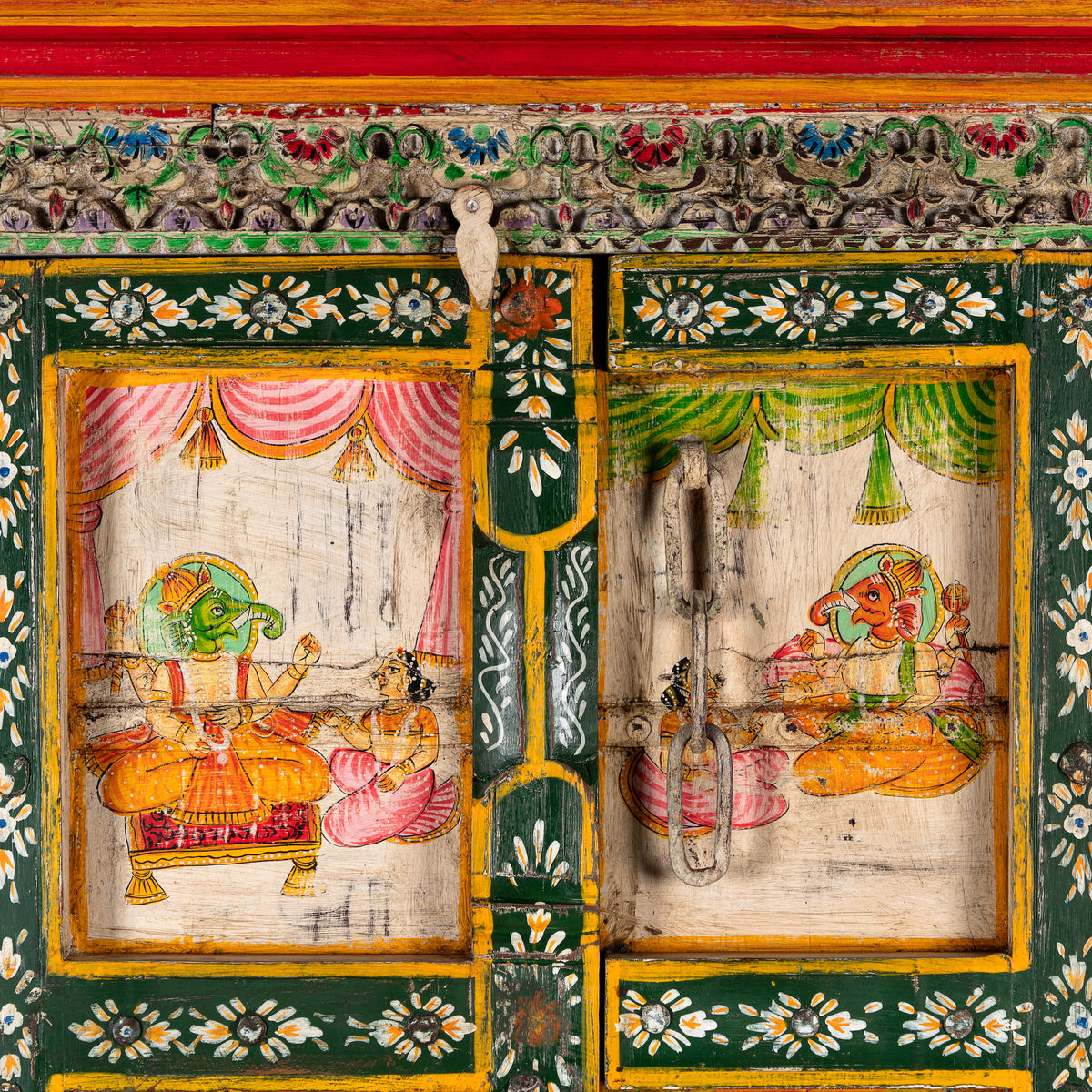 Close up image of a a tall, hand-painted Indian wooden cabinet with colorful traditional illustrations, set in a bright, modern living room next to a large potted plant.