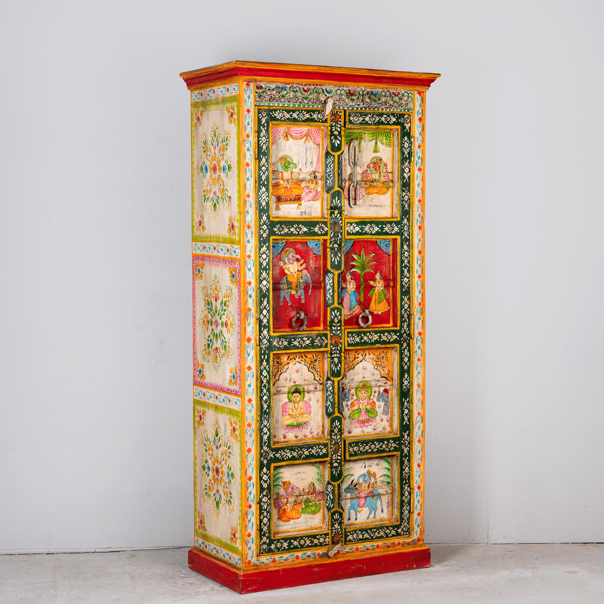 Side angle image of a a tall, hand-painted Indian wooden cabinet with colorful traditional illustrations, set in a bright, modern living room next to a large potted plant.