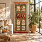 Lifestyle image of a tall, hand-painted Indian wooden cabinet with colorful traditional illustrations, set in a bright, modern living room next to a large potted plant.