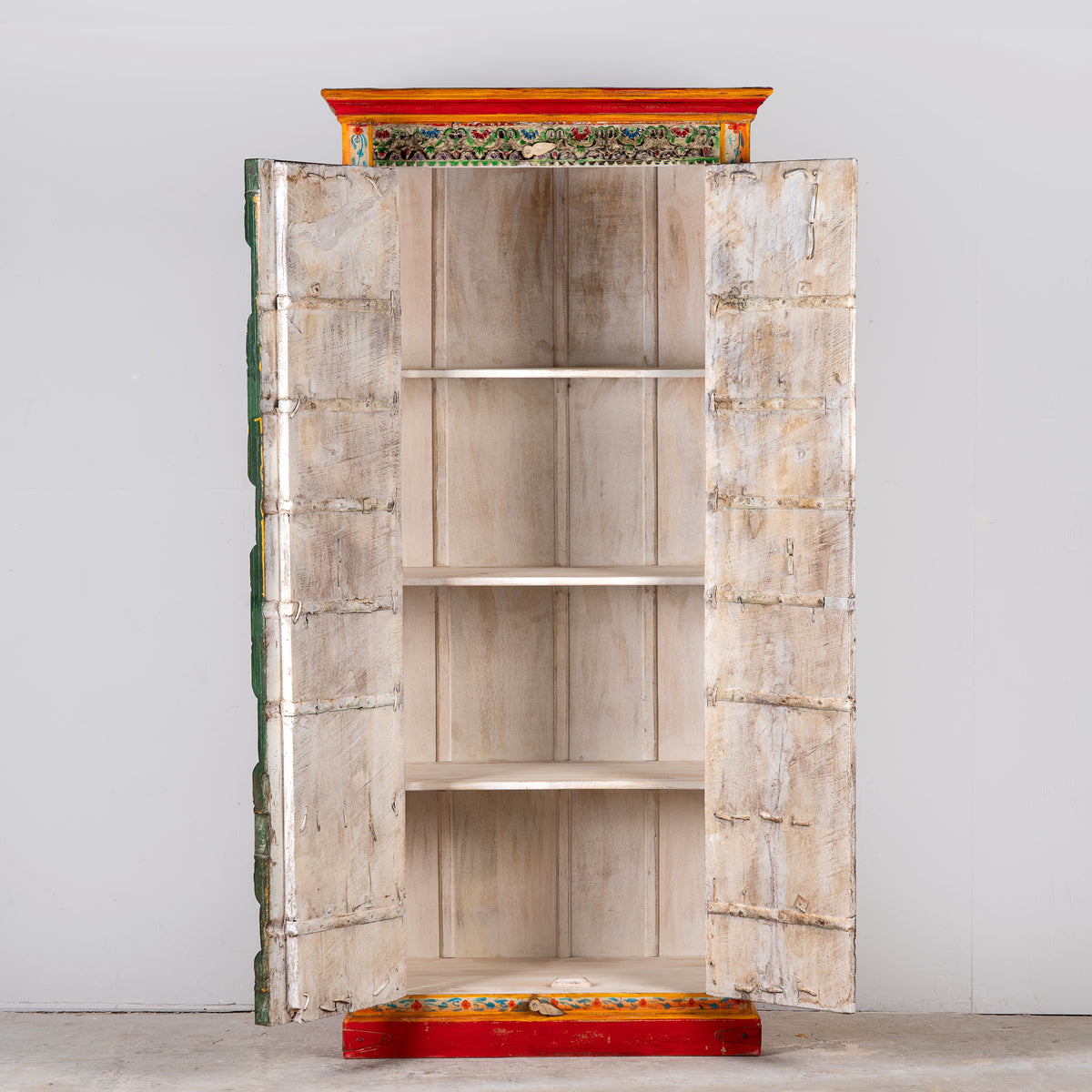 Image showing open door view of a a tall, hand-painted Indian wooden cabinet with colourful traditional illustrations, set in a bright, modern living room next to a large potted plant.
