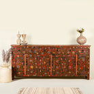 A hand-painted reclaimed wood sideboard with intricate floral designs in vibrant colors. The sideboard has multiple compartments and is positioned on a striped rug against a plain wall.