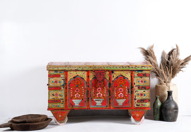 A hand-painted teak wood storage chest on wheels, featuring vibrant red, yellow, and green patterns with floral and geometric designs, displayed against a white background with a few decorative items around it.