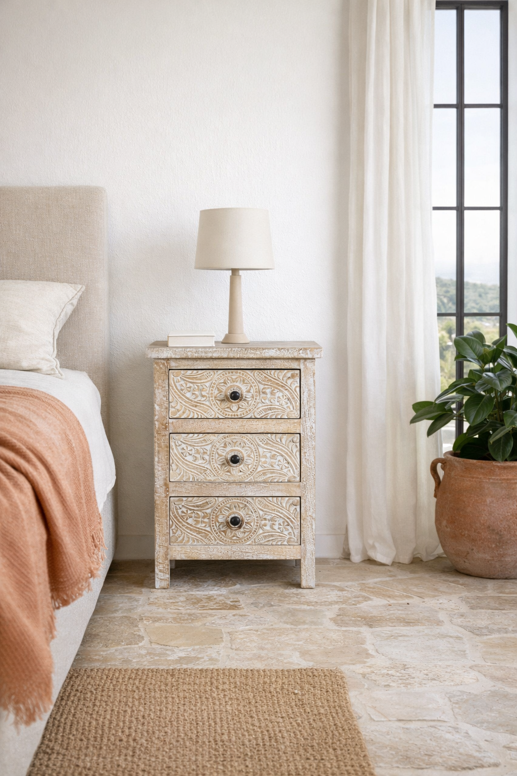 Lifestyle imagination of a pair of mango wood nightstands Ved in a whitewashed vintage finish, featuring hand-carved drawer fronts with ornate circular and leafy motifs. Stylish and compact vintage-style bedside tables with three drawers for elegant storage.