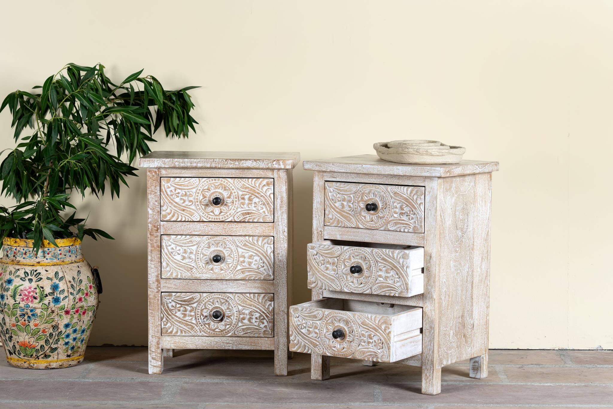 Side angle image with open drawers of a pair of mango wood nightstands Ved in a whitewashed vintage finish, featuring hand-carved drawer fronts with ornate circular and leafy motifs. Stylish and compact vintage-style bedside tables with three drawers for elegant storage.