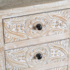 Close up image of a pair of mango wood nightstands Ved in a whitewashed vintage finish, featuring hand-carved drawer fronts with ornate circular and leafy motifs. Stylish and compact vintage-style bedside tables with three drawers for elegant storage.