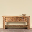 A rustic carved wooden console table with a distressed finish, featuring multiple drawer fronts with brass-tone studs and geometric inlays, a lower open shelf, and carved side panels. It is shown against a plain beige wall on a brick floor with a small decorative bowl on top.