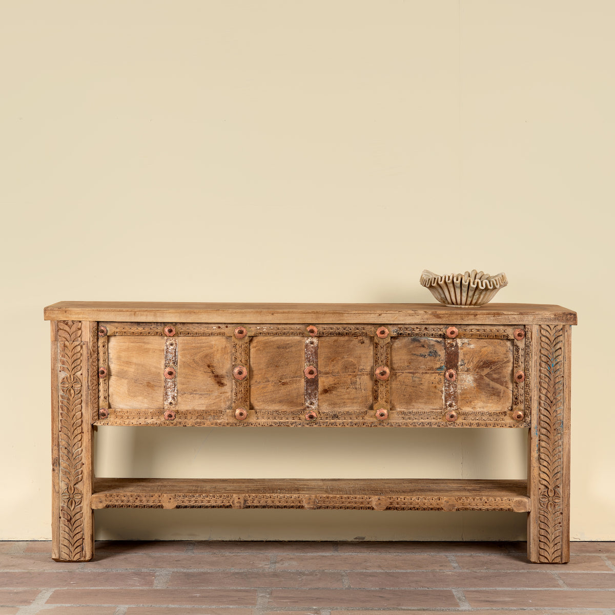 A rustic carved wooden console table with a distressed finish, featuring multiple drawer fronts with brass-tone studs and geometric inlays, a lower open shelf, and carved side panels. It is shown against a plain beige wall on a brick floor with a small decorative bowl on top.