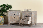 Side angle image of a pair of hand-carved mango wood nightstands Vikram with three drawers each, showcasing intricate floral motifs in a distressed vintage-style finish. Elegant and functional bedside tables with timeless artisan appeal.