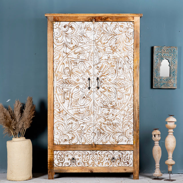Hand-carved mango wood wardrobe with whitewashed floral carvings and a natural wood body, featuring three doors and a drawer, displayed in a room setting with decorative items around it.