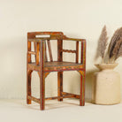 Side angle image of a hand-painted armchair crafted from reclaimed teak wood, featuring intricate traditional patterns in earthy tones on the backrest, seat, and frame, with a rustic and vintage finish.
