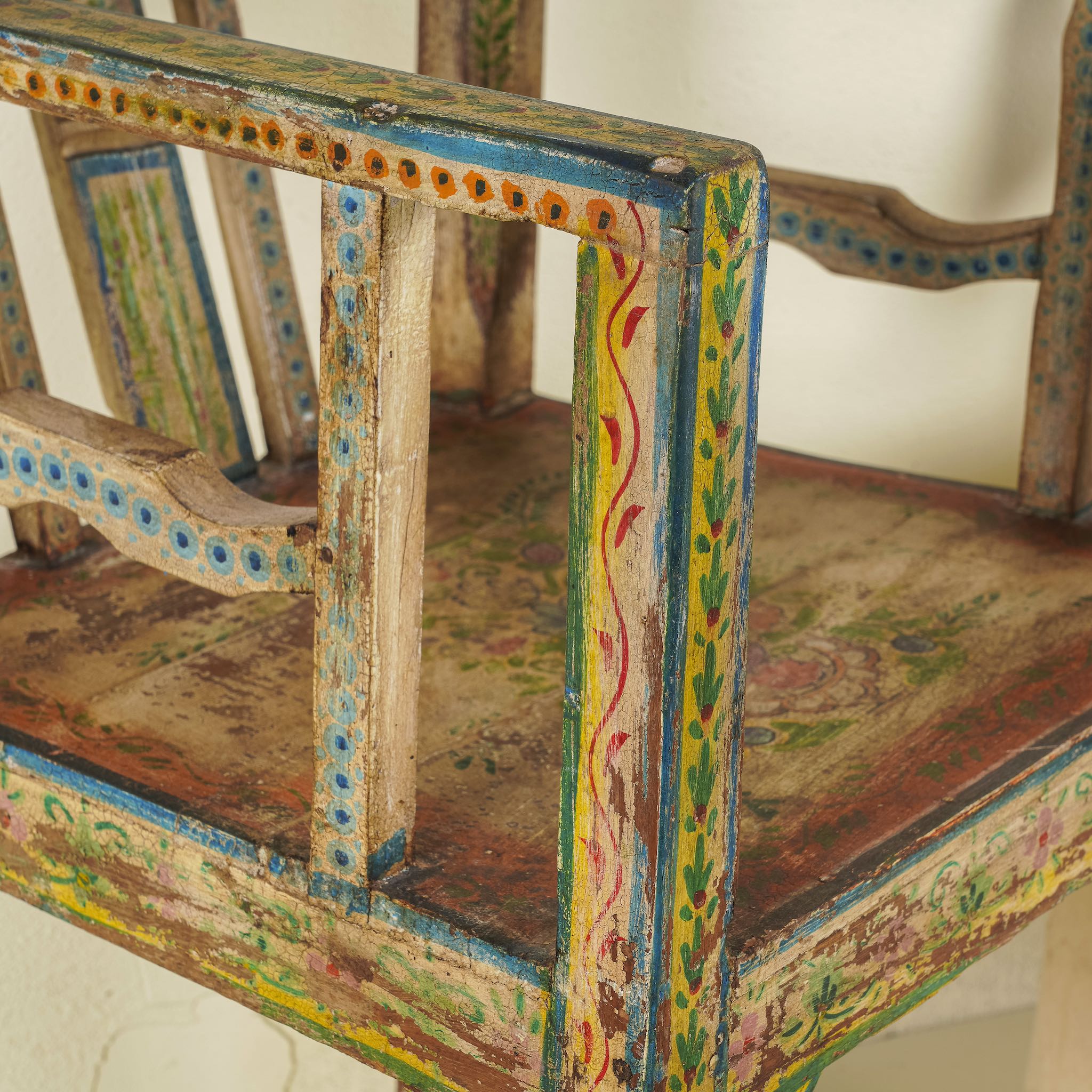 Arm rest of a hand-painted armchair crafted from reclaimed teak wood, featuring intricate traditional patterns in earthy tones on the backrest, seat, and frame, with a rustic and vintage finish.

