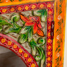 Close up picture of a hand-painted showcase cabinet made from mango wood, featuring a circular glass-front display, floral motifs in rich orange, green, and red tones, and five drawers for added storage.

