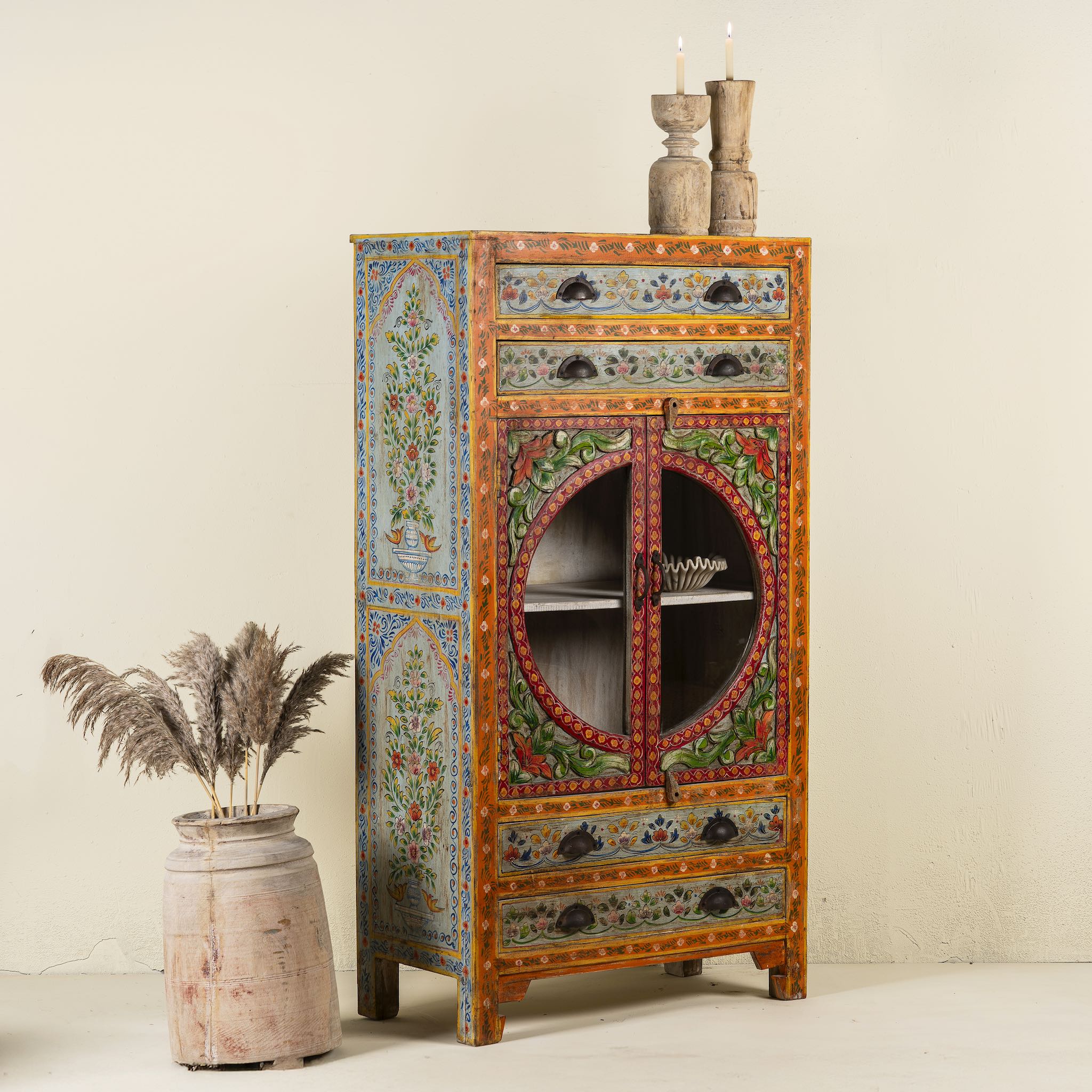 Side angle picture of a hand-painted showcase cabinet made from mango wood, featuring a circular glass-front display, floral motifs in rich orange, green, and red tones, and five drawers for added storage.

