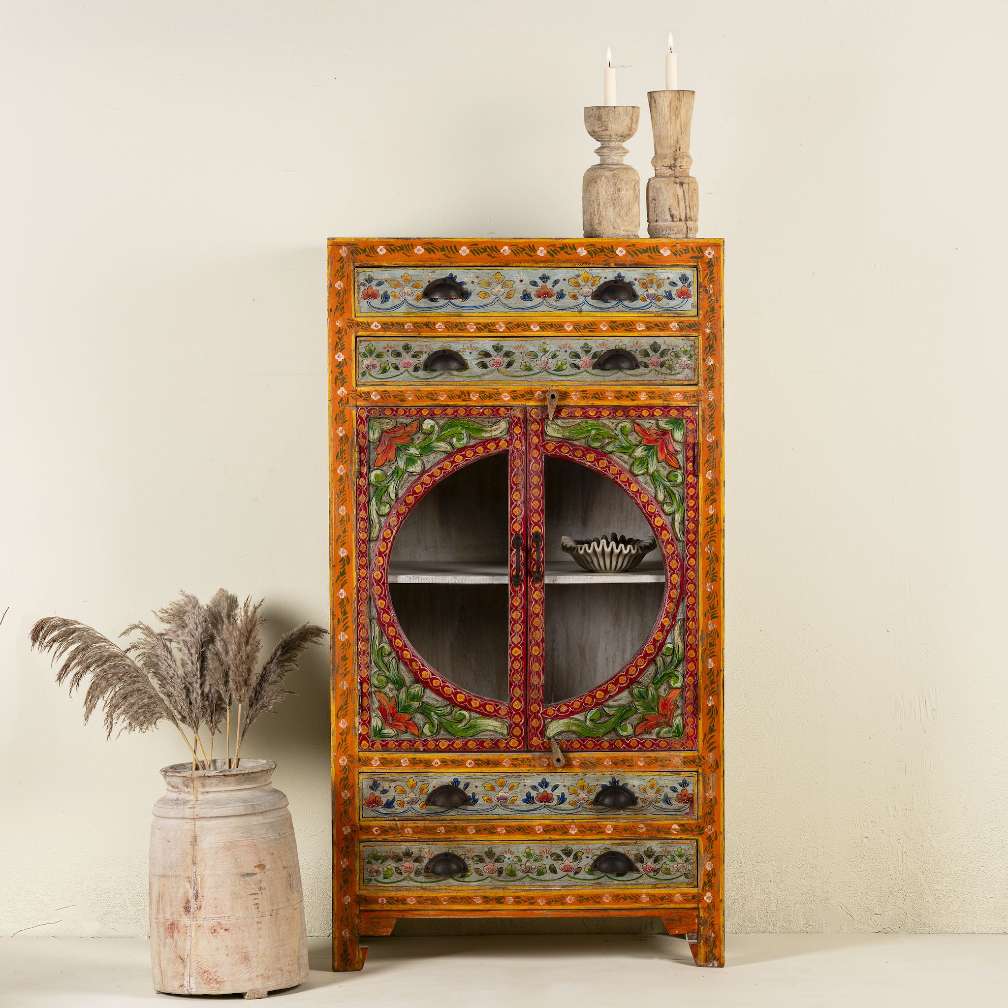Hand-painted showcase cabinet made from mango wood, featuring a circular glass-front display, floral motifs in rich orange, green, and red tones, and five drawers for added storage.

