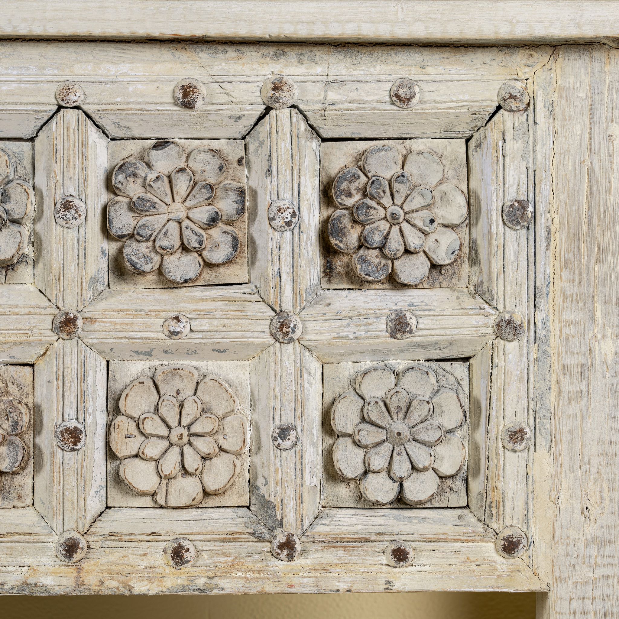 Close up image of a hand carved console table made from reclaimed teak wood, featuring a detailed front panel with geometric floral motifs and a distressed whitewashed finish, complemented by a lower shelf for added storage or display.
