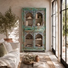 Lifesyle image of a hand-carved mango wood showcase with glass doors and drawers – carved wooden furniture in distressed turquoise and blue, combining artisan detail with vintage charm.