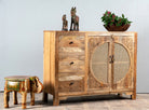 Side angle image of a hand carved mango wood cabinet in natural finish with 3 drawers and 2 rattan doors.